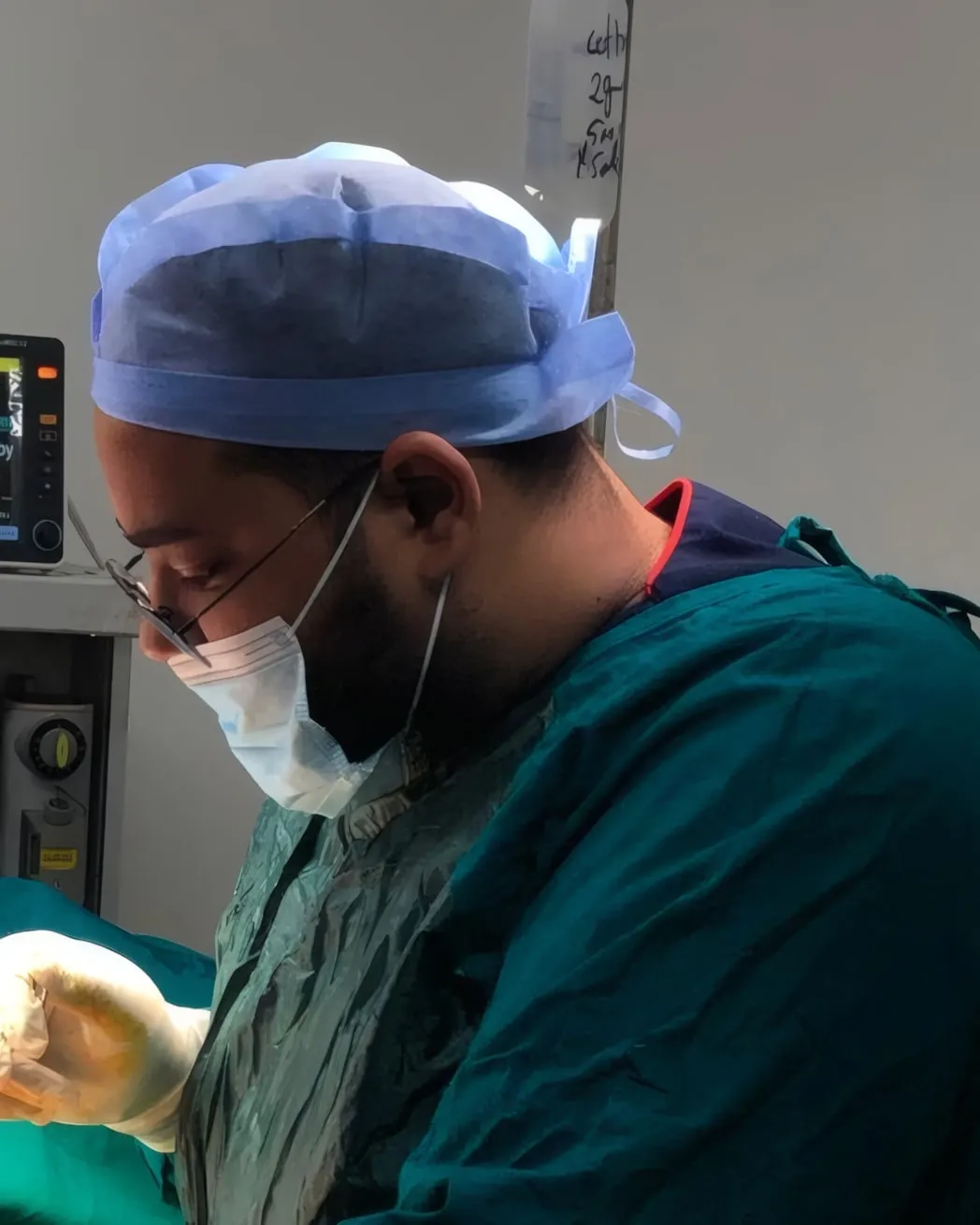 د. أبانوب أيمن بزي جراحي أخضر وغطاء رأس ونظارة وقناع، منهمكًا في عملية مع الفريق الجراحي، وخلفه شاشة متابعة المريض